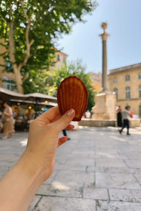 A hand holding up a madeleine in front of a square in Aix-en-Provence