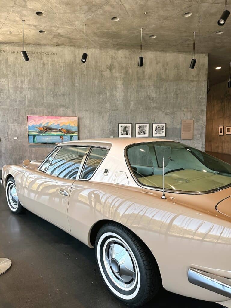 The interior of one of the LACMA galleries. There is a beige vintage car, and art pieces hanging on the concrete wall in the background.
