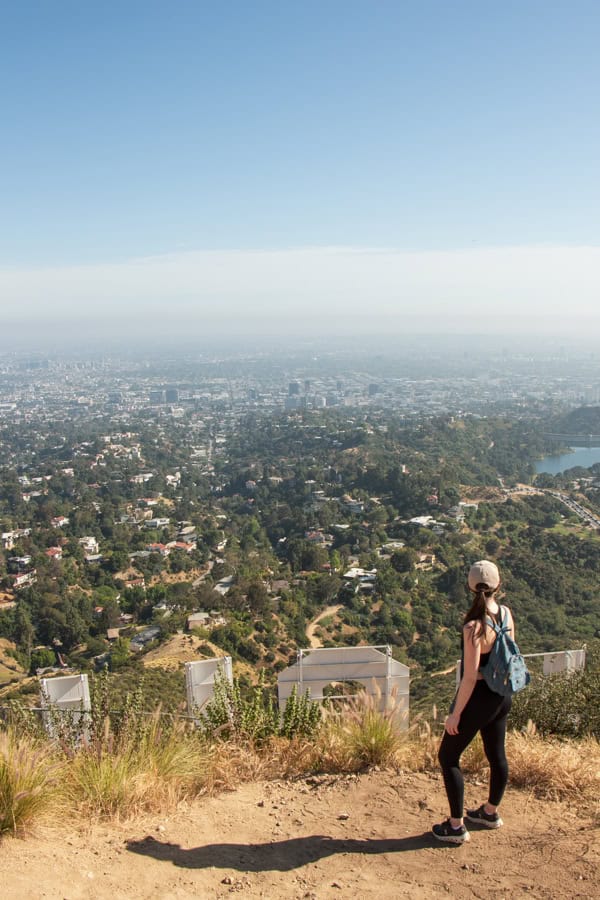 A woman standing behind the Hollywood Sign, looking at the view of the Hollywood Reservoir and Los Angeles. You can see the very top of the first few letters of the Hollywood Sign.