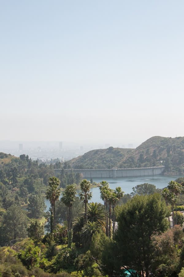 The view of Los Angeles from Lake Hollywood Park. There are palm trees in front of the Hollywood Reservoir, and you can faintly see buildings behind haze in the distance.