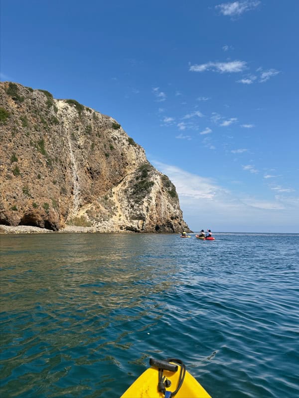 The tip of a yellow kayak in the water at Scorpion Anchorage. There are kayakers in the distance