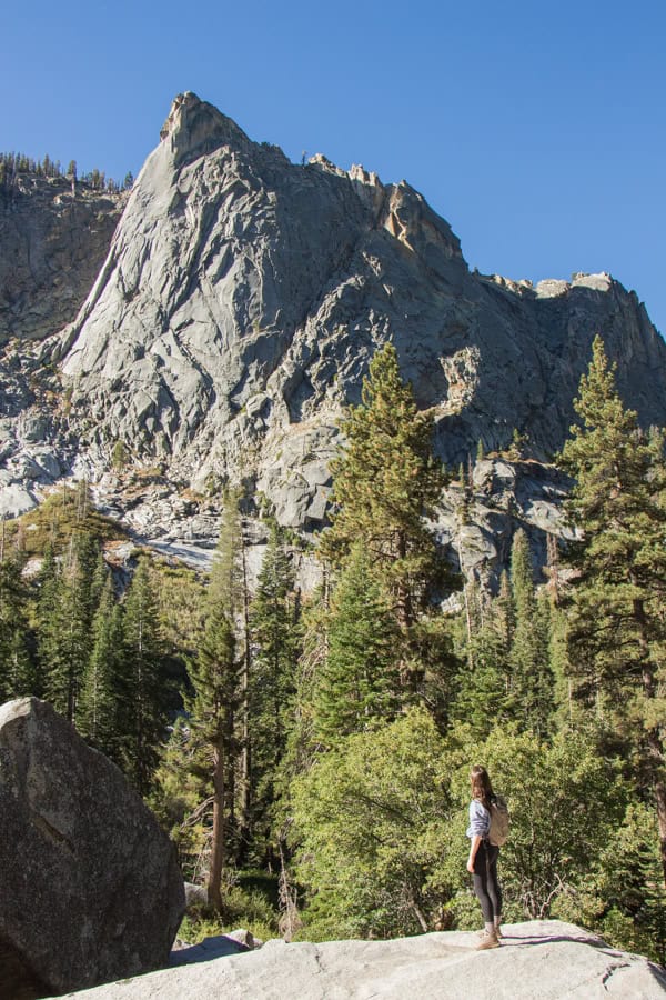 A woman standing on a boulder looking up at the rock face above the Tokopah Falls Trail. There are trees in the valley below.